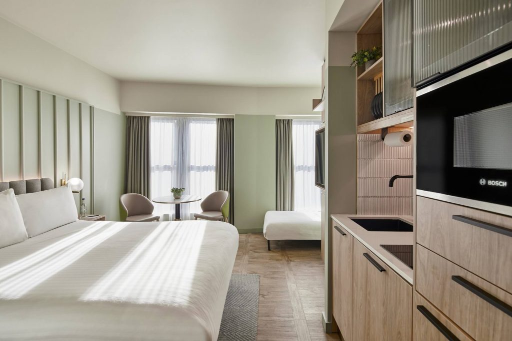 A master St. Paul’s studio featuring a bed with a green throw, a round table and two chairs by a window, and a kitchenette with sink and built-in microwave.