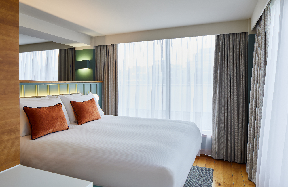 Bright bedroom at master Farringdon London serviced apartments, featuring a large double bed with crisp white linens and two orange accent pillows, framed by floor-to-ceiling windows and soft natural light.