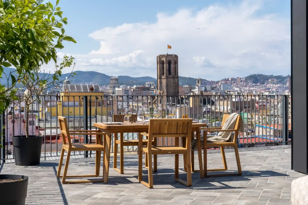 Outdoor dining table for four on a spacious terrace overlooking Barcelona’s rooftops and historic bell tower.