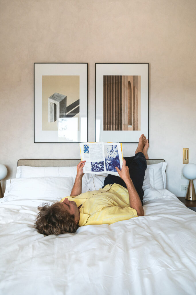 Man relaxing on a bed reading a book, in a cozy bedroom with framed artwork above the headboard.