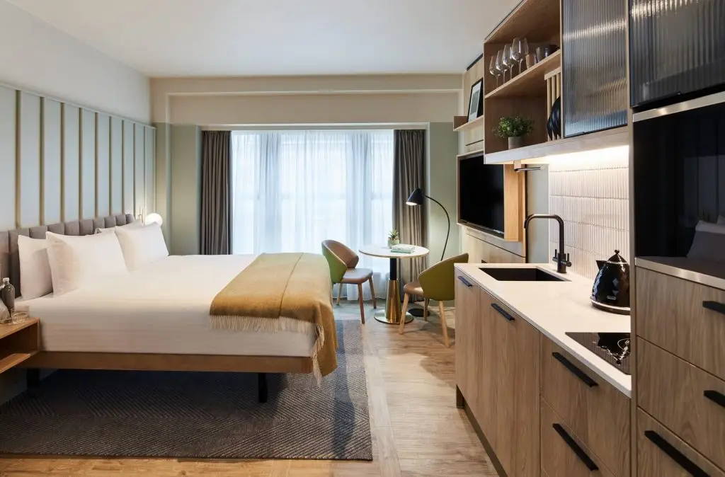 A master St. Paul’s studio with a king bed, small round table and two chairs by the windows, and a kitchenette with wood cabinetry.