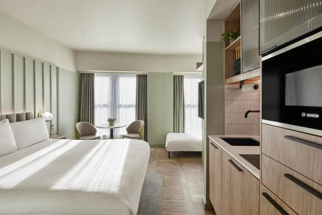 A master St. Paul’s studio featuring a bed with a green throw, a round table and two chairs by a window, and a kitchenette with sink and built-in microwave.