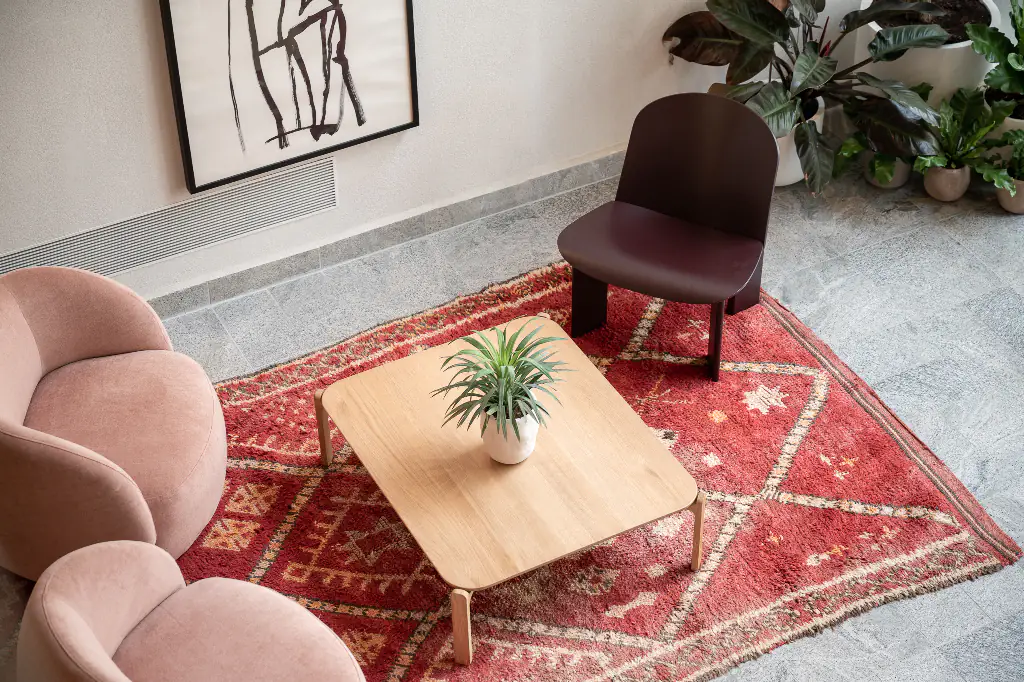 Chic sitting area at master Mazeh with soft pink armchairs, a burgundy accent chair, wooden coffee table, and a red patterned rug, surrounded by potted plants and abstract wall art.