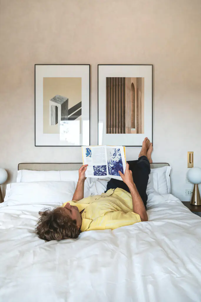 Man relaxing on a bed reading a book, in a cozy bedroom with framed artwork above the headboard.